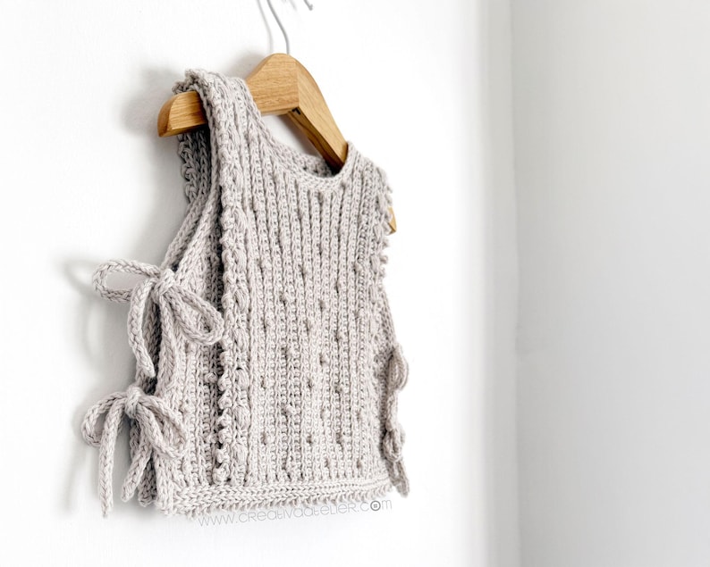 Peut inclure: Un haut sans manches en crochet gris clair avec un motif textur&eacute; et des liens lat&eacute;raux. Le haut est suspendu &agrave; un cintre en bois contre un mur blanc. Les liens lat&eacute;raux sont nou&eacute;s en n&oelig;uds.