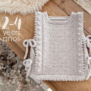 Può includere: Un gilet lavorato a maglia grigio chiaro per bambini, con dettagli a pallini e lacci laterali. Il gilet ha uno scollo tondo e un motivo strutturato. Il testo "2-4 years años" è visibile.