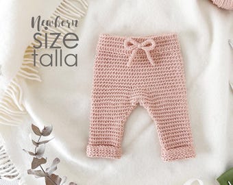Puede incluir: Un pantalón de bebé de color rosa tejido a crochet con cintura de cordón. El pantalón está colocado sobre una superficie blanca. El texto "Newborn size talla" es visible en la esquina superior izquierda de la imagen.