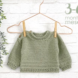 May include: A sage green knitted baby sweater with wooden buttons hanging on a copper hanger. The sweater is for 3-6 months old babies.