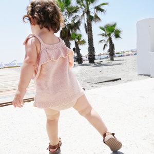 Op de afbeelding: Een jong kind draagt een roze gebreide jurk met een gerimpelde halslijn en bruine sandalen en loopt op een zandstrand met palmbomen op de achtergrond.
