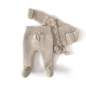 May include: A beige knitted two-piece set consisting of a long-sleeved cardigan with small pom-pom accents and a pair of footed leggings featuring a ribbed waistband with a decorative drawstring tie and small pom-poms on the feet.