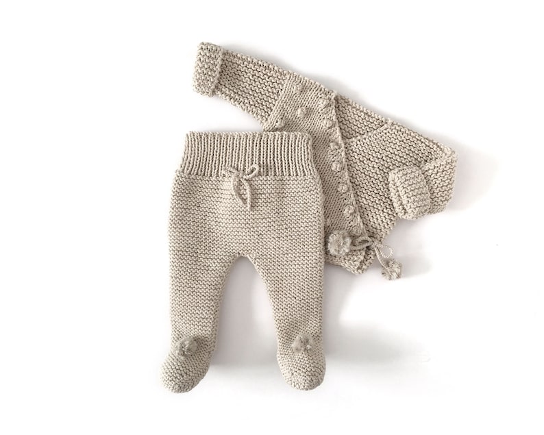 May include: A beige knitted two-piece set consisting of a long-sleeved cardigan with small pom-pom accents and a pair of footed leggings featuring a ribbed waistband with a decorative drawstring tie and small pom-poms on the feet.