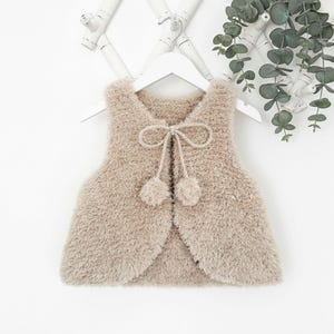 Op de afbeelding: Een beige, pluizig vest met een strik en twee pompons. Het vest hangt aan een witte hanger tegen een witte achtergrond. Het vest is gemaakt van een zacht, getextureerd materiaal. Het is een kinderkledingstuk.