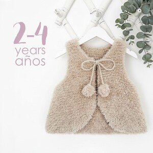 Op de afbeelding: Een beige, pluizig vest voor kinderen, hangend aan een witte hanger. Het vest heeft een strik en twee pompons aan de voorkant. De tekst "2-4 years años" staat linksboven. De achtergrond is wit.