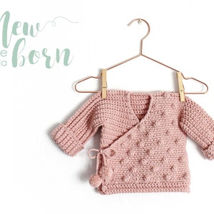 Könnte beinhalten: Ein rosa gehäkelter Baby-Pullover mit Wickel-Stil und einem Bommel-Detail. Der Pullover hängt an einem Kupfer-Kleiderbügel mit Holz-Wäscheklammern. Der Text "New born size talla" ist in Grün auf weißem Hintergrund geschrieben.