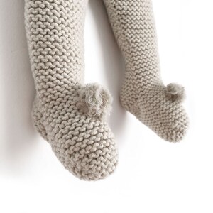 May include: A pair of beige knit leggings with integrated footies, featuring a garter stitch texture throughout the legs and a small, decorative pom-pom detail attached to the top of each foot area, providing a soft and textured appearance.