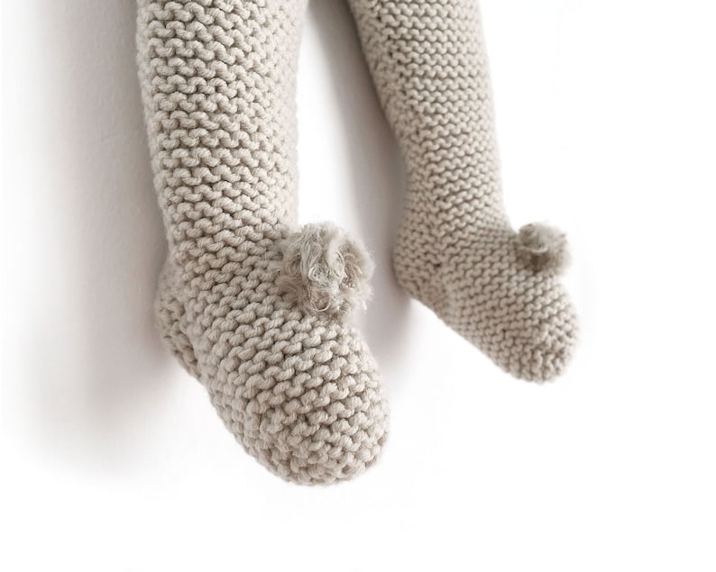 May include: A pair of beige knit leggings with integrated footies, featuring a garter stitch texture throughout the legs and a small, decorative pom-pom detail attached to the top of each foot area, providing a soft and textured appearance.