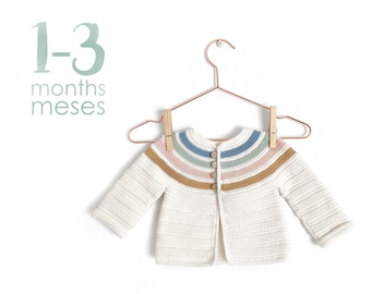 Peut inclure: Un cardigan bébé en crochet blanc avec des rayures arc-en-ciel et une fermeture boutonnée. Le cardigan est suspendu à un cintre en cuivre avec des pinces à linge en bois. Le texte "1-3 mois meses" est écrit en vert sur un fond blanc.