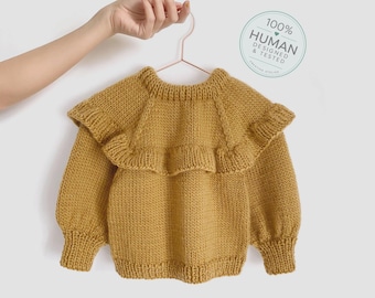May include: A mustard yellow long-sleeve knit sweater featuring a wide ruffled collar, ribbed cuffs, and a ribbed neckline. A circular badge in the upper right corner displays the text 100% HUMAN DESIGNED & TESTED CREATIVE ATELIER.