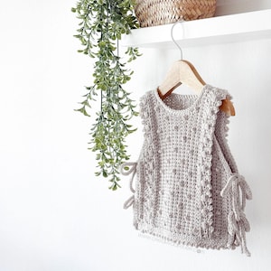 Peut inclure: Un haut sans manches en crochet gris clair avec un motif textur&eacute; et des liens d&eacute;coratifs sur les c&ocirc;t&eacute;s. Le haut est suspendu &agrave; un cintre en bois sur fond blanc. Le haut mesure environ 30 cm de long.