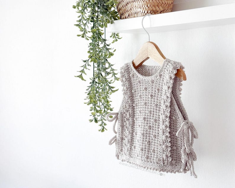 Peut inclure: Un haut sans manches en crochet gris clair avec un motif textur&eacute; et des liens d&eacute;coratifs sur les c&ocirc;t&eacute;s. Le haut est suspendu &agrave; un cintre en bois sur fond blanc. Le haut mesure environ 30 cm de long.