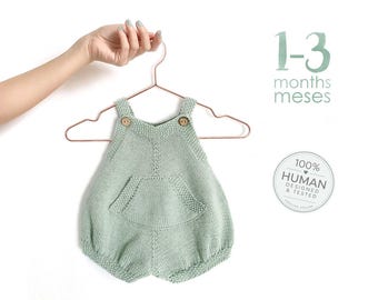 May include: A sage green knitted romper featuring a central pocket, two small wooden buttons at the shoulder straps, and a textured knit pattern. The text 1-3 months meses and a circular badge stating 100% HUMAN DESIGNED & TESTED are visible.