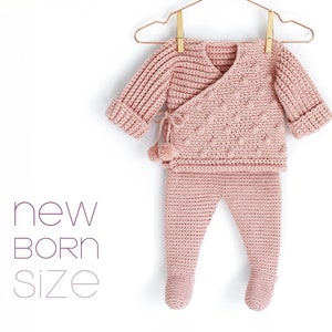 Può includere: Un completo per neonato in crochet rosa con una maglia a fascia e pantaloni abbinati. La maglia ha un design testurizzato e un dettaglio a pompon. Il completo è etichettato "new born size".