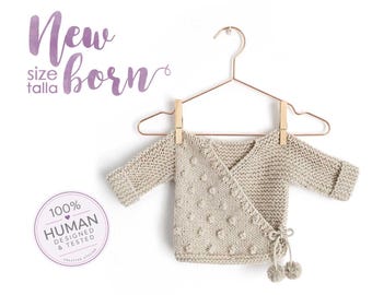 May include: A beige knit wrap sweater for a newborn featuring a textured bobble pattern on the front, long sleeves with cuffs, and a side tie closure with two small pom-poms. The text New born size talla and 100% HUMAN DESIGNED & TESTED appears.