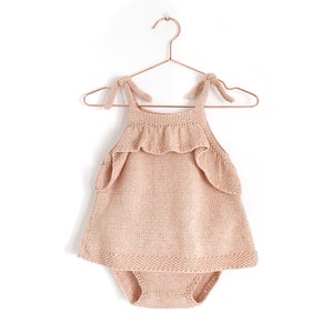 May include: A pale pink knitted baby dress with a ruffled neckline and straps that tie at the shoulders. The dress is hanging on a copper hanger.
