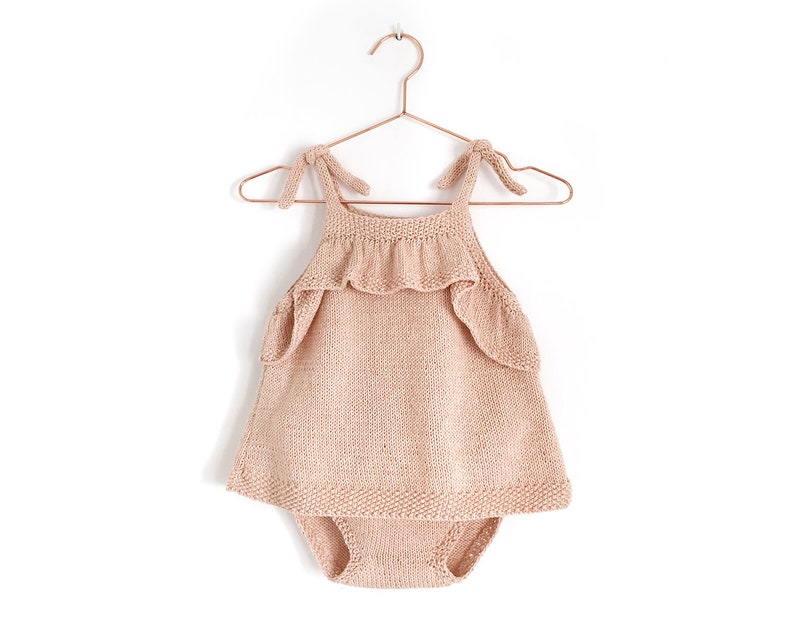 May include: A pale pink knitted baby dress with a ruffled neckline and straps that tie at the shoulders. The dress is hanging on a copper hanger.