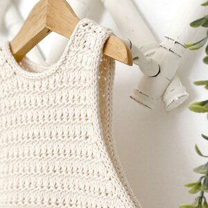 May include: A cream-colored crocheted sleeveless top hanging on a wooden hanger. The top has a simple, open-stitch design.