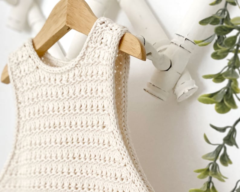May include: A cream-colored crocheted sleeveless top hanging on a wooden hanger. The top has a simple, open-stitch design.