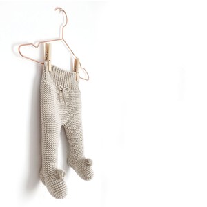 May include: Beige knit leggings with a ribbed waistband, a decorative drawstring tie, and integrated footed sections featuring small pom-poms at the toes. The garment is suspended from a copper-coloured wire hanger by two wooden clothespins.