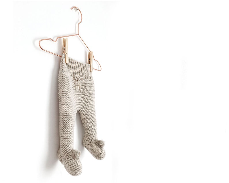 May include: Beige knit leggings with a ribbed waistband, a decorative drawstring tie, and integrated footed sections featuring small pom-poms at the toes. The garment is suspended from a copper-coloured wire hanger by two wooden clothespins.