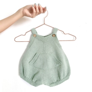 Könnte beinhalten: Ein salbeigrüner gestrickter Baby-Strampler mit einer Tasche und Knopfverschlüssen. Der Strampler hängt an einem Kupferdraht-Kleiderbügel.