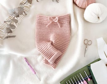 Puede incluir: Un pantalón de bebé de crochet rosa con cintura de cordón. El pantalón está sobre una superficie blanca con un gancho de crochet y lana.