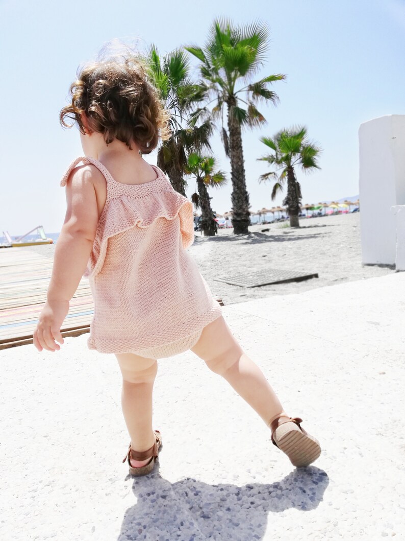 May include: A young child wearing a pink knitted dress with a ruffled neckline and brown sandals walks on a sandy beach with palm trees in the background.