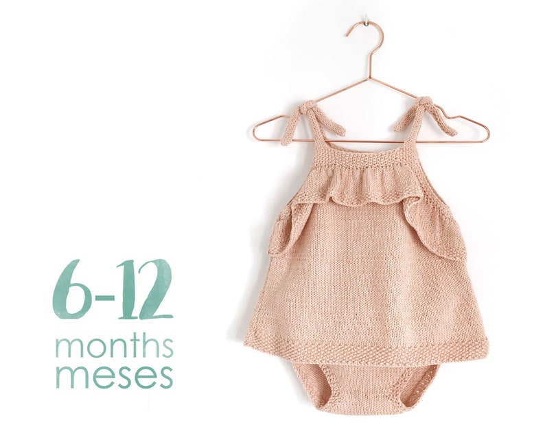Op de afbeelding: Een gebreide baby romper met een geruit randje en bandjes. De romper is lichtroze van kleur en hangt aan een koperen hanger. De tekst "6-12 maanden meses" is onder de romper gedrukt.