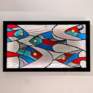 Könnte beinhalten: Abstrakte Glasmalerei mit einem Design aus wirbelnden Formen in Blautönen, Rot und Gelb. Das Kunstwerk ist in Schwarz gerahmt.