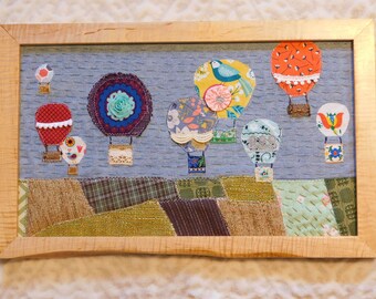 May include: A framed artwork featuring a blue background with colorful hot air balloons made of fabric. The balloons are decorated with various patterns and designs, including flowers, birds, and geometric shapes. The artwork is framed in a natural wood frame.