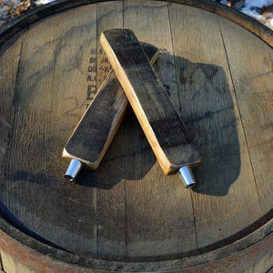 May include: Two wooden beer tap handles with silver spouts. The handles are made from reclaimed wood and are laying on top of a wooden barrel.