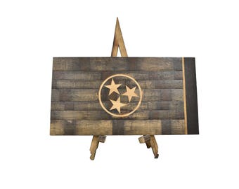 May include: A wooden sign featuring the Tennessee state flag, displayed on a wooden easel. The sign has a dark, weathered appearance with the state's three stars emblem in the center. The sign is approximately 24 inches wide.