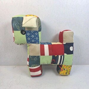 May include: A stuffed animal shaped like a dog, constructed from a patchwork of various fabric squares. The patterns include red and white stripes, navy blue with white polka dots, floral prints, and solid light green, tan, and red sections.