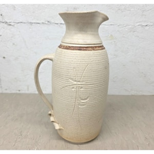 May include: A tall, off-white ceramic pitcher with a textured, ribbed surface and a single handle. The body features a carved, abstract linear design and a brown glazed band around the neck. The base has a small, decorative curled clay accent.