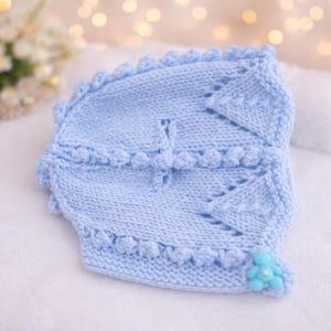 May include: A light blue knitted baby bonnet with a scalloped edge and a small blue flower accent. The bonnet is made of soft yarn and features a delicate design, perfect for a newborn. The bonnet is approximately 8 inches in diameter.