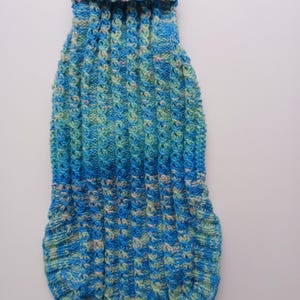 May include: A hand-knitted dog sweater in shades of blue and green. The sweater features a ribbed collar and a textured stitch pattern throughout the body. A small tag is attached near the bottom. The sweater is designed to keep a small dog warm.
