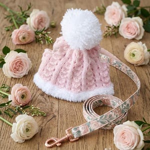 May include: A pink knitted dog hat with a white pom-pom and trim, paired with a floral patterned leash. The leash has a rose gold clasp. The items are arranged on a wooden surface with scattered pink and white roses.