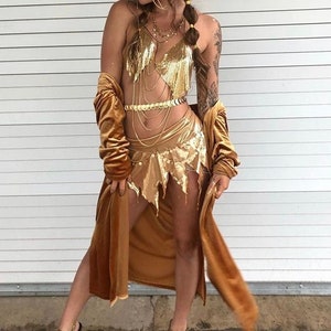 Peut inclure: Une femme portant un haut et une jupe en sequins dorés avec une ceinture à chaîne dorée et un couvre-chef doré. Elle porte également une robe en velours marron et des talons compensés dorés.