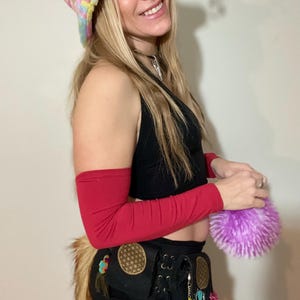 May include: A vibrant tie-dye bucket hat, black crop top, red arm sleeves, and a black utility belt with decorative charms. A fluffy purple ball is held in the hand. A faux fur tail is attached to the belt.