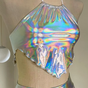 Peut inclure: Haut de bikini argenté holographique et jupe assortie. Le haut est de forme triangulaire avec un bord festonné et une bretelle blanche. La jupe est taille haute. Le tissu reflète un arc-en-ciel de couleurs.