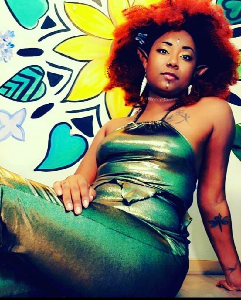 May include: A woman with red hair and elf ears is wearing a green and gold metallic halter top and matching pants. She is sitting in front of a colorful wall with a mandala design.