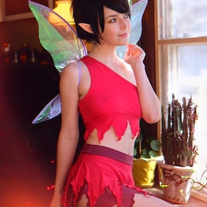 May include: A two-piece red fairy costume featuring a one-shoulder crop top with a jagged hem and a matching skirt with a layered, pointed hemline. The outfit includes large, iridescent, translucent wings with a subtle purple tint.