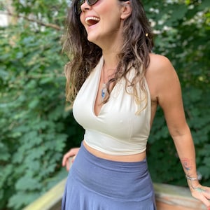 May include: A cream-colored halter top with a deep V-neckline and an open back design, paired with a solid slate blue skirt featuring a wide, high-rise waistband. The outfit is worn with dark-tinted sunglasses and a silver pendant necklace.