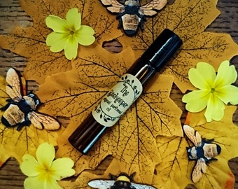 May include: A brown glass bottle with a label that reads "The Beekeeper Original Perfume Oil". The bottle is surrounded by yellow flowers and black and yellow bees on a brown wooden background.