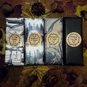 May include: Four different scented soy wax melts, each with a unique design and label. The labels feature text describing the scent, such as "Ye Olde Library", "Vanilla Sandalwood & Calness", "Weedsmoke & Leather", and "Crackling Log Fire".