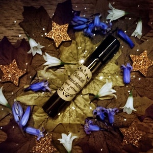 Może przedstawiać: Mała szklana butelka z czarną zakrętką i brązową etykietą z napisem "Celestial Incantations Original Perfumed Oil". Butelka jest otoczona niebieskimi i białymi kwiatami, złotym brokatem i brązowymi liśćmi.