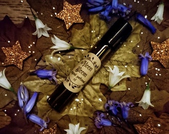 May include: A small brown glass bottle with a label that reads "Alceon Bloom,  spiral perfume oil". The bottle is surrounded by blue and white flowers, gold glitter stars, and brown leaves.