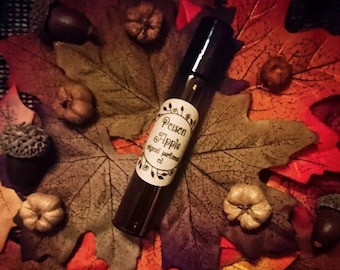 Puede incluir: Una botella de vidrio marrón con una tapa negra y una etiqueta blanca que dice "Poison Apple Original Perfume Oil" sobre un lecho de hojas de otoño.