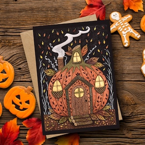 May include: A Halloween-themed greeting card featuring a pumpkin house illustration with a smoking chimney. The card is accompanied by pumpkin, ghost, and skeleton-shaped cookies, and autumn leaves on a wooden surface.